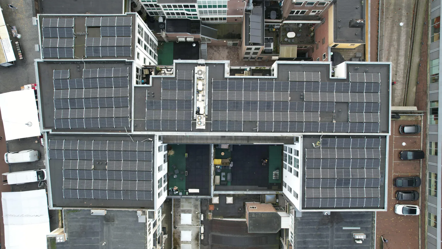 Luchtfoto recht van boven op een stedelijk bouwblok waarvan alle platte daken volledig zijn volgelegd met zonnepanelen, met binnenplaatsen, dakopbouwen en geparkeerde auto's langs de randen