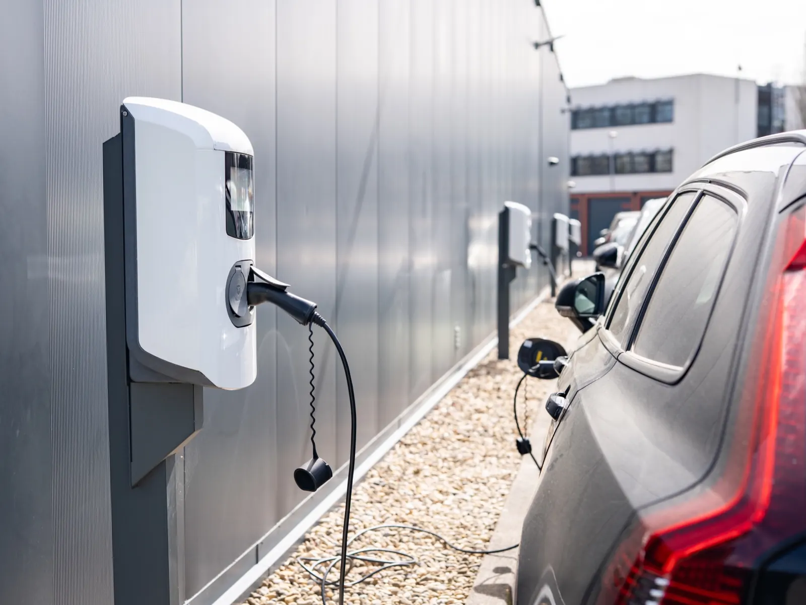 Witte AC-laadpunten aan de gevel van een bedrijfspand, met laadkabels naar auto's die op de grindstrook ernaast staan