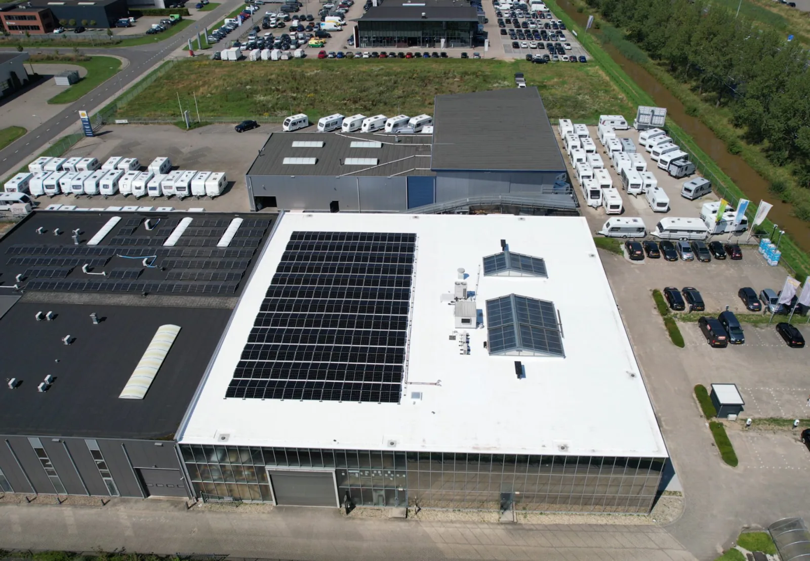 Luchtfoto van een bedrijventerrein waar op een deel van het witte platte dak zonnepanelen liggen, met campers en auto's op het terrein eromheen