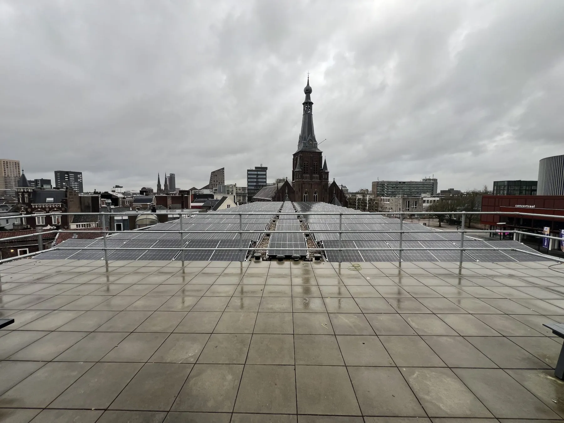 Uitzicht over het betegelde platte dak van een wooncomplex met zonnepanelen op lage stellages, met daarachter de stad en een kerktoren
