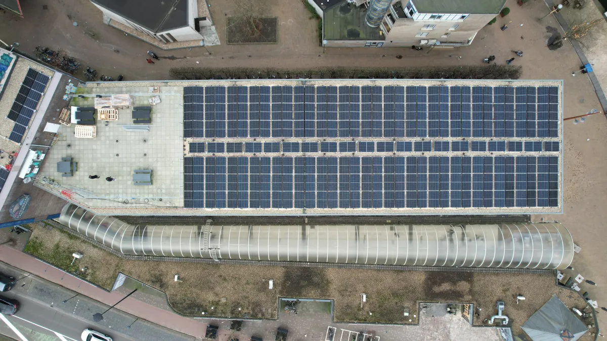 Luchtfoto recht boven een langgerekt dak in het centrum van Tilburg dat over vrijwel de hele lengte met zonnepanelen is belegd, met een glazen passage ernaast