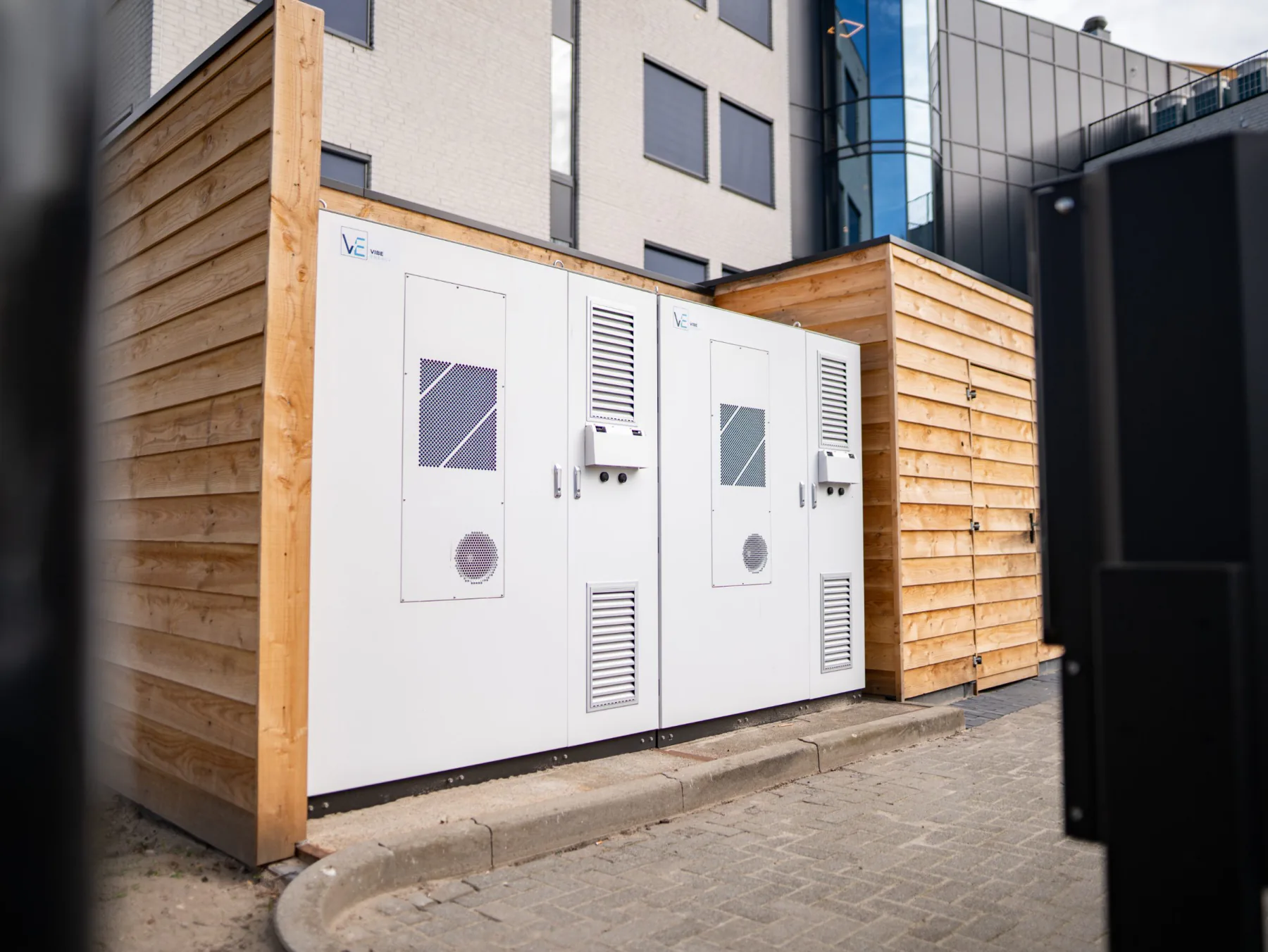 Twee witte batterijkasten van Vibe Energy in een houten omkasting op de bestrating naast een bedrijfspand