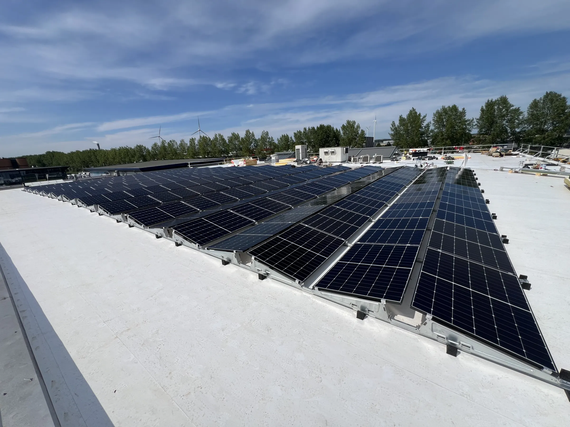 Rijen zonnepanelen op het witte platte dak van een bedrijfspand, met bomen en windturbines aan de horizon